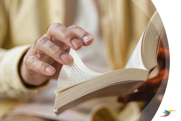 Imagem da mão de uma pessoa idosa folheando um livro. Na lateral direita, detalhes brancos e a logo do Colibri.